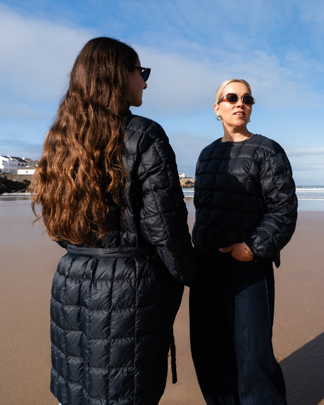 Two women in black down coats  standing on a beach with a clear blue sky.