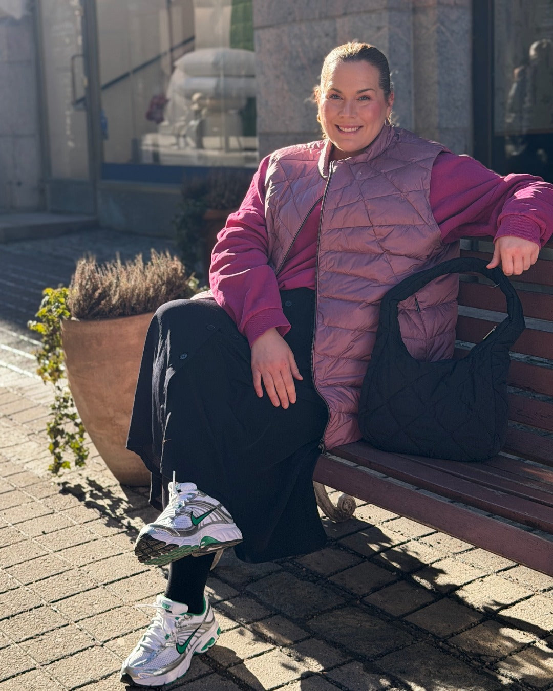 Woman sitting on a bench wearing a pink down vest and black pants, with a plant and building in the background.