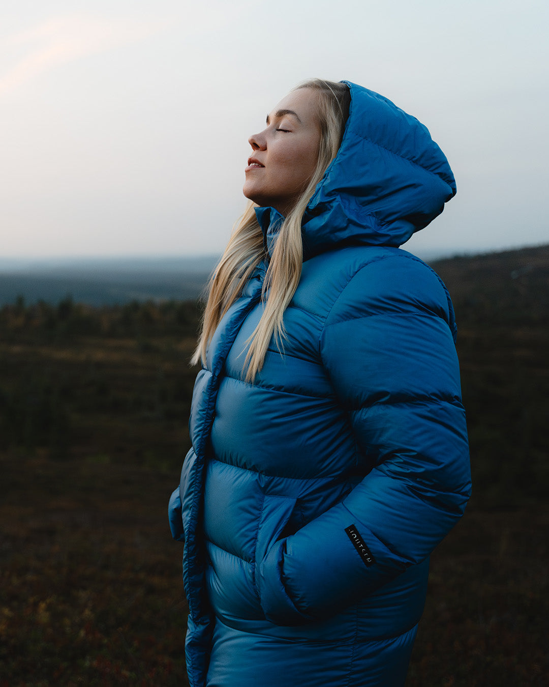 A blonde woman stands in a forested landscape wearing a blue (color: Blue butterfly) down coat with a small Joutsen logo on the sleeve. She has the hood up and her hands in the coat pockets. Her face is tilted slightly upward with her eyes closed.