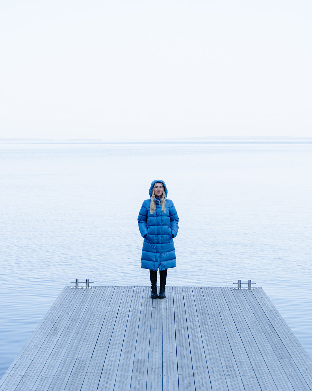 A blonde woman stands on a pier wearing a blue (color: Blue butterfly) knee-length down coat with horizontal quilting. A foggy body of water is visible in the background. She has her hands in the coat pockets and is wearing the coat’s attached hood.