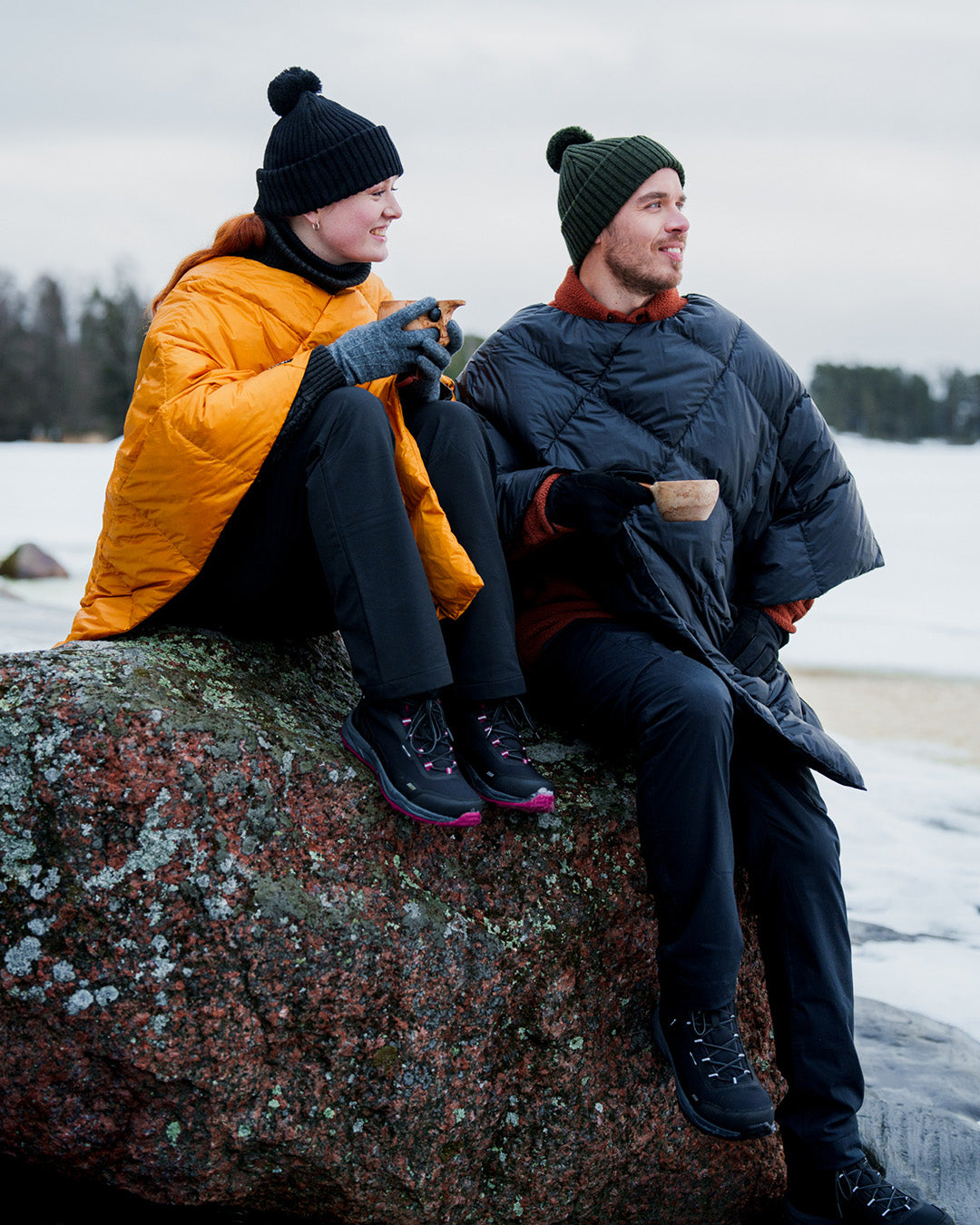 Two people are sitting on a large rock in a winter landscape, wearing diamond-quilted Otava down ponchos. The person on the left is wearing a yellow poncho (color: amber), and the one on the right a dark one (color: charcoal black). Both are wearing beanies and holding drinks.