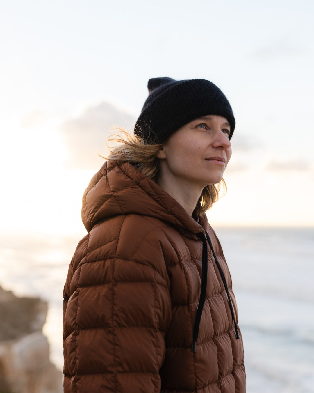 Person wearing a brown down hoodie and black beanie standing by a body of water.