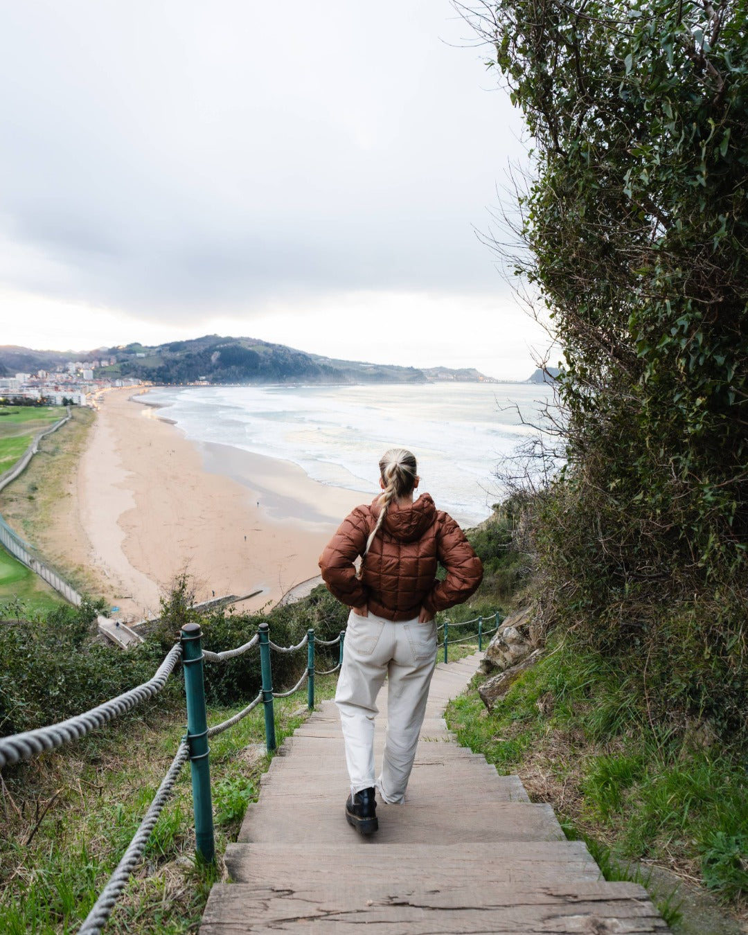 Person walking up a wooden staircase towards a beach with a scenic view. She is wearing a brown down hoodie.