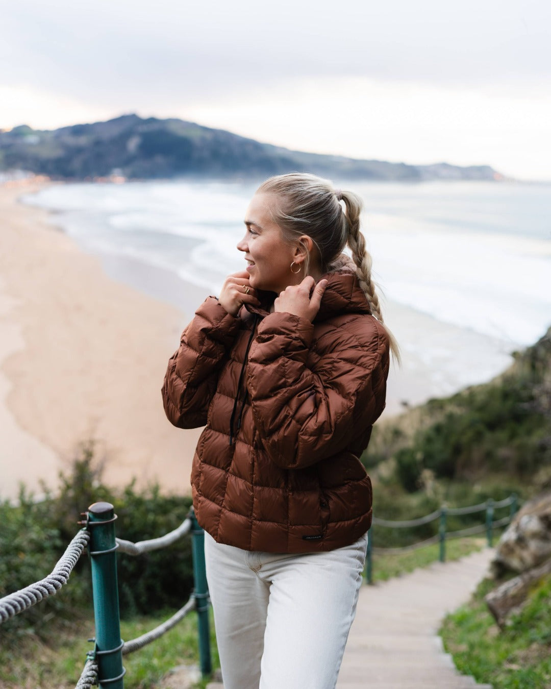 Woman in a brown down hoodie standing on a coastal path with a scenic view.