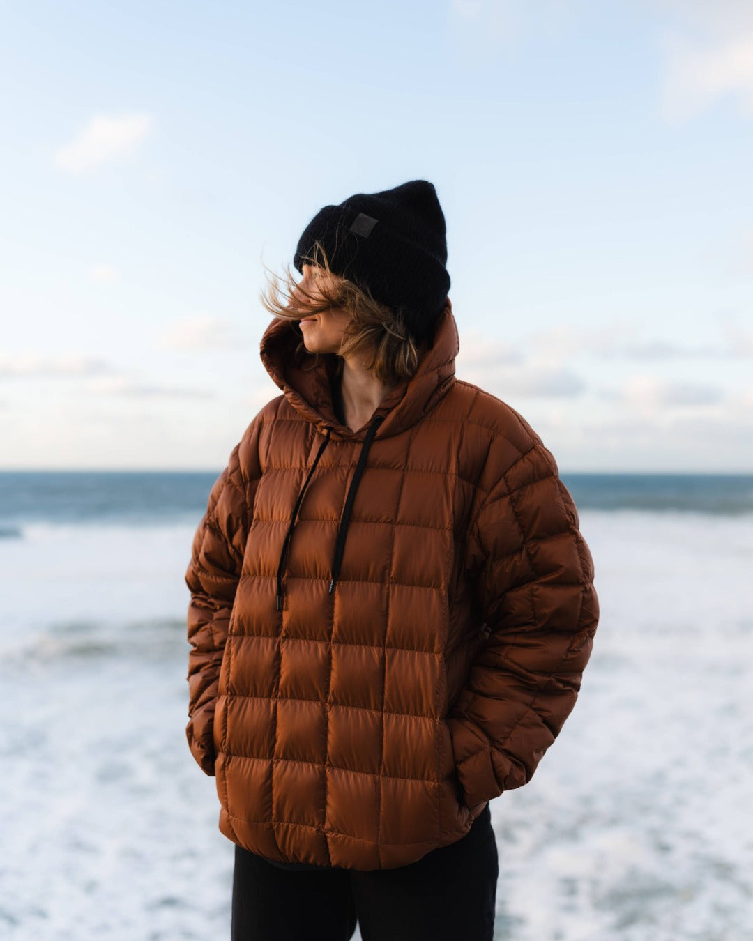 Person wearing a brown down hoodie and black beanie standing on a beach.