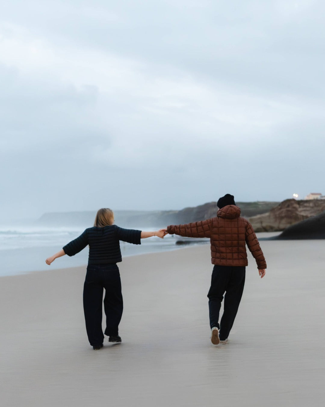 Two people walking hand in hand on a beach with a cloudy sky. The other one is wearing ja black down bolero and the other one is wearing a brown down hoodie.