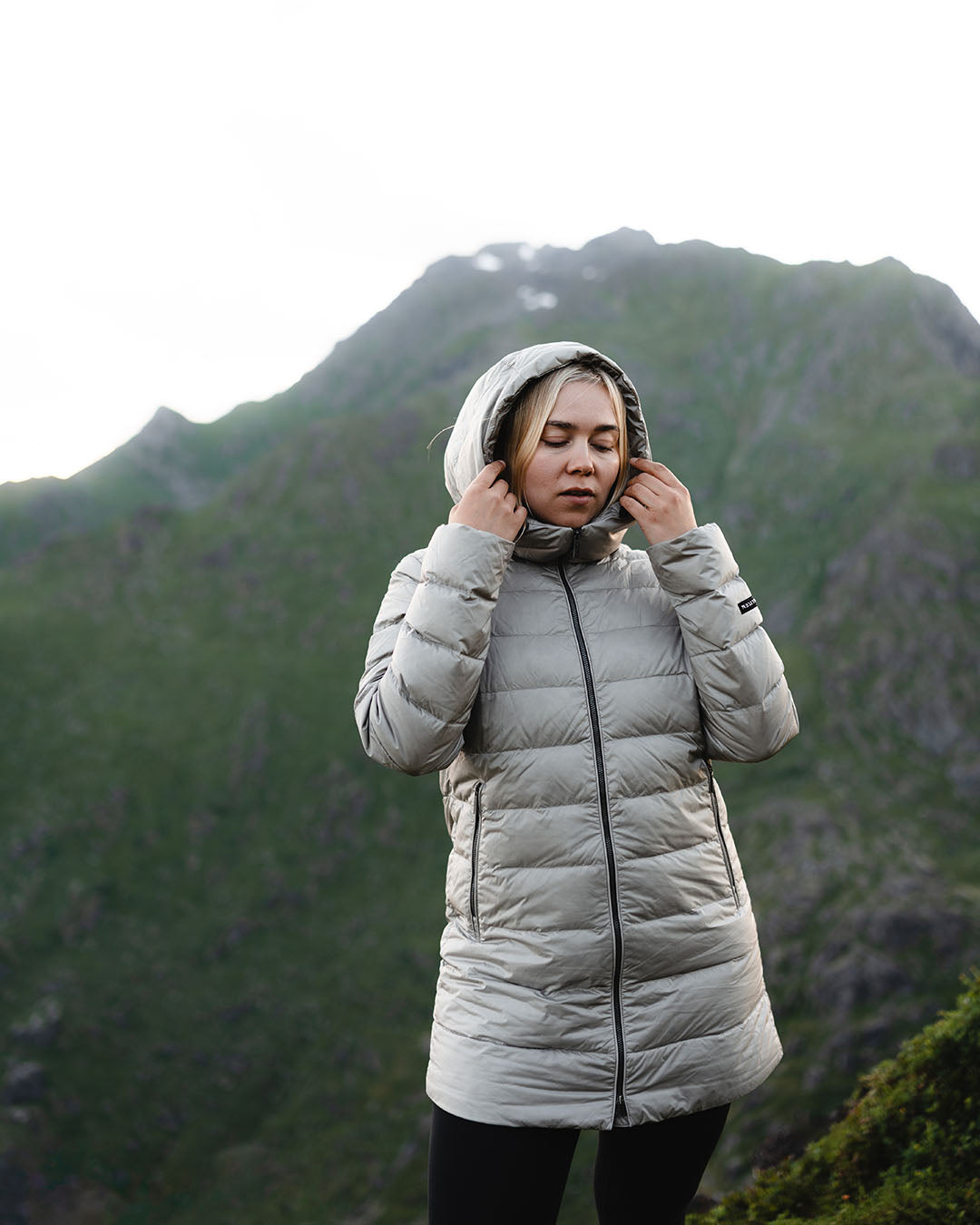 A person stands in a mountainous landscape wearing a beige Ruut light down coat (color: champagne) with the hood up, holding the edges of the hood.