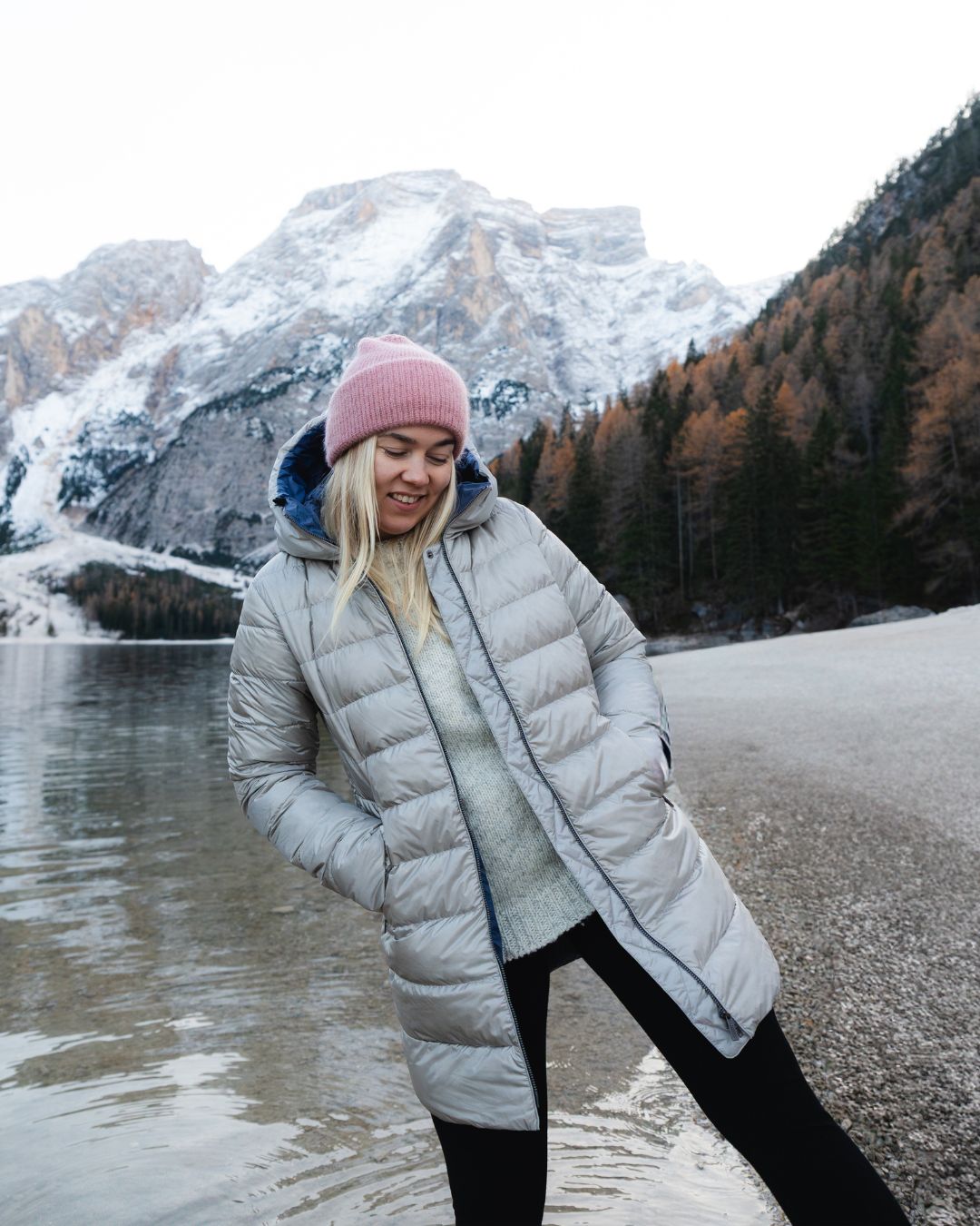Person wearing a grey coat and pink beanie standing by a snowy mountain lake.