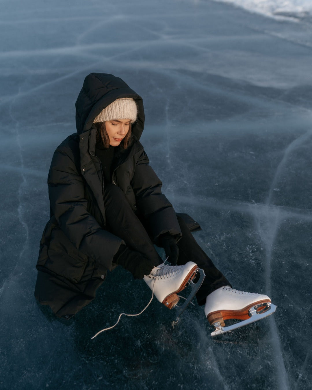 Person sitting on a frozen lake with ice skates on. They are wearing a black Siiri down coat.