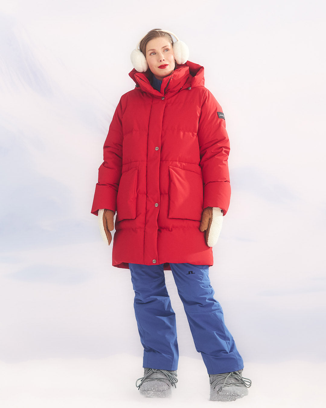 A woman is standing in a winter landscape dressed in a red (color: chili pepper) Siiri down parka coat. She is wearing large white earmuffs, blue insulated pants, and grey winter boots.