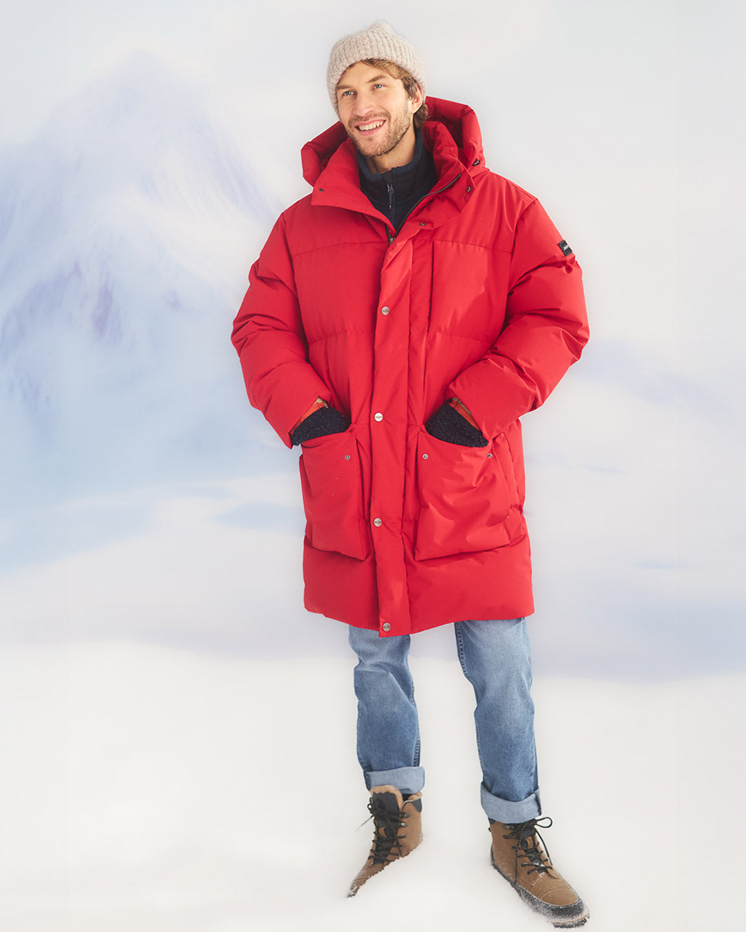 A man is standing in a snowy landscape wearing a long, red (color: chili pepper) Tuisku down parka coat with a large hood and front pockets. He is also wearing blue jeans, a light grey knitted beanie, and brown lace-up winter boots.
