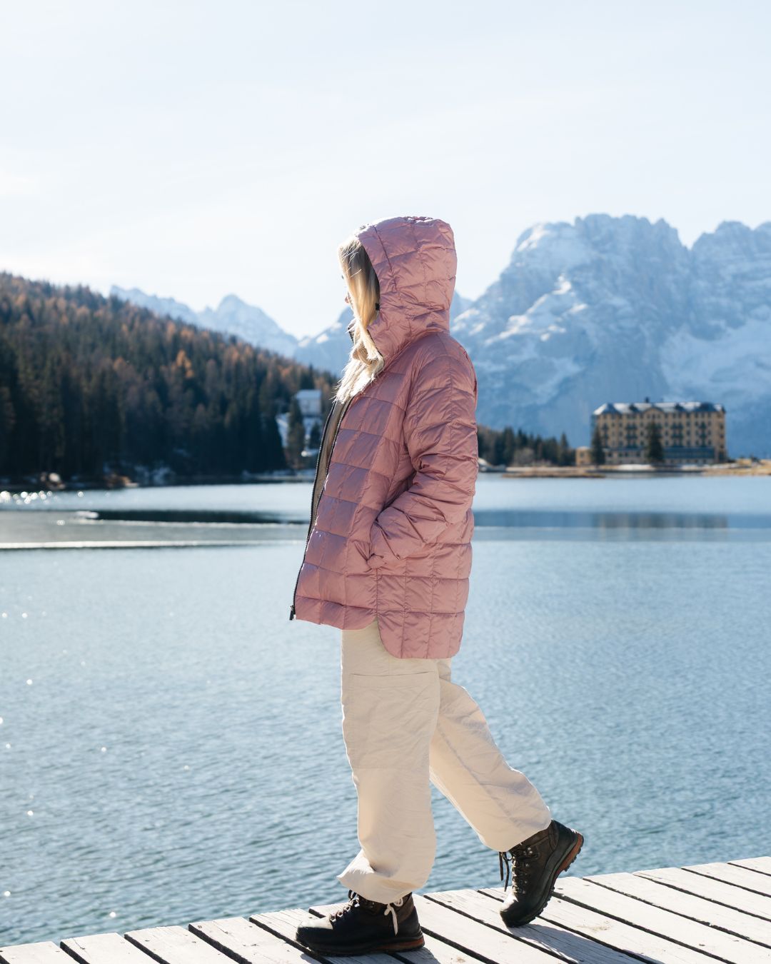 Person wearing a pink hooded light down coat standing on a dock by a lake with mountains in the background