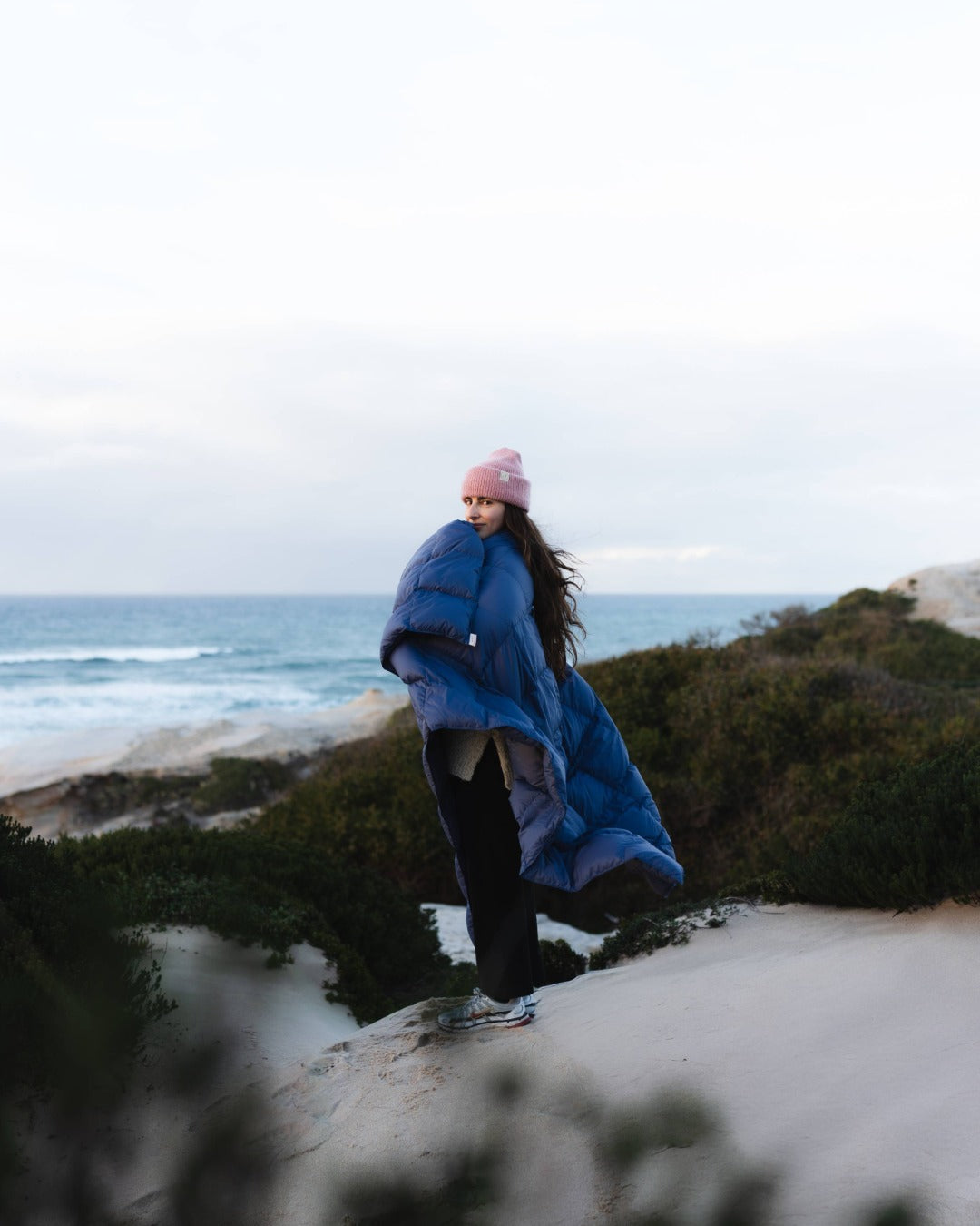 Person wrapped in a blue blanket standing on a beach with ocean and sky in the background