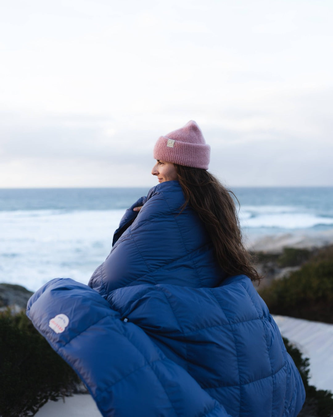 Person is wrapped in blue down blanket and pink beanie sitting on a rocky outcrop overlooking the ocean.