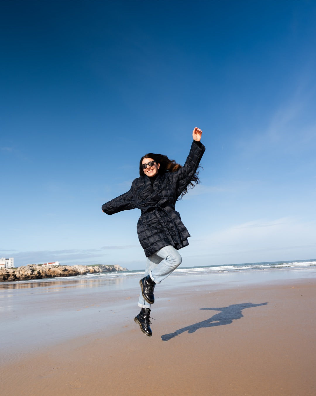Person jumping on a beach wearing a black down jacket.