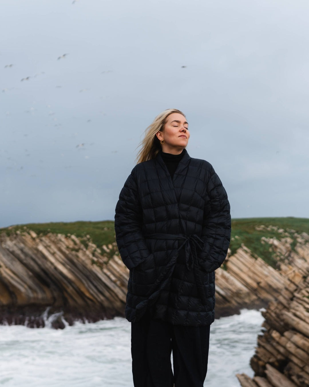 Person wearing a black down jacket standing on a coastal landscape with wooden structures and a cloudy sky.