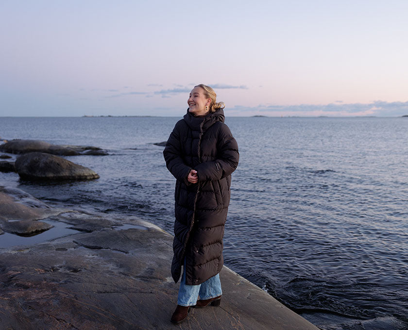A woman standing on a rocky shoreline by the ocean, wearing a long dark down coat.