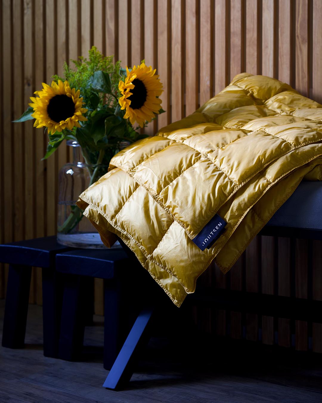 A quilted yellow (color: Tuscan sun) Kulkuri down blanket is folded on a dark bench, next to a glass vase filled with sunflowers and green leaves. In the background, a light wooden paneled wall creates a warm and cozy atmosphere.