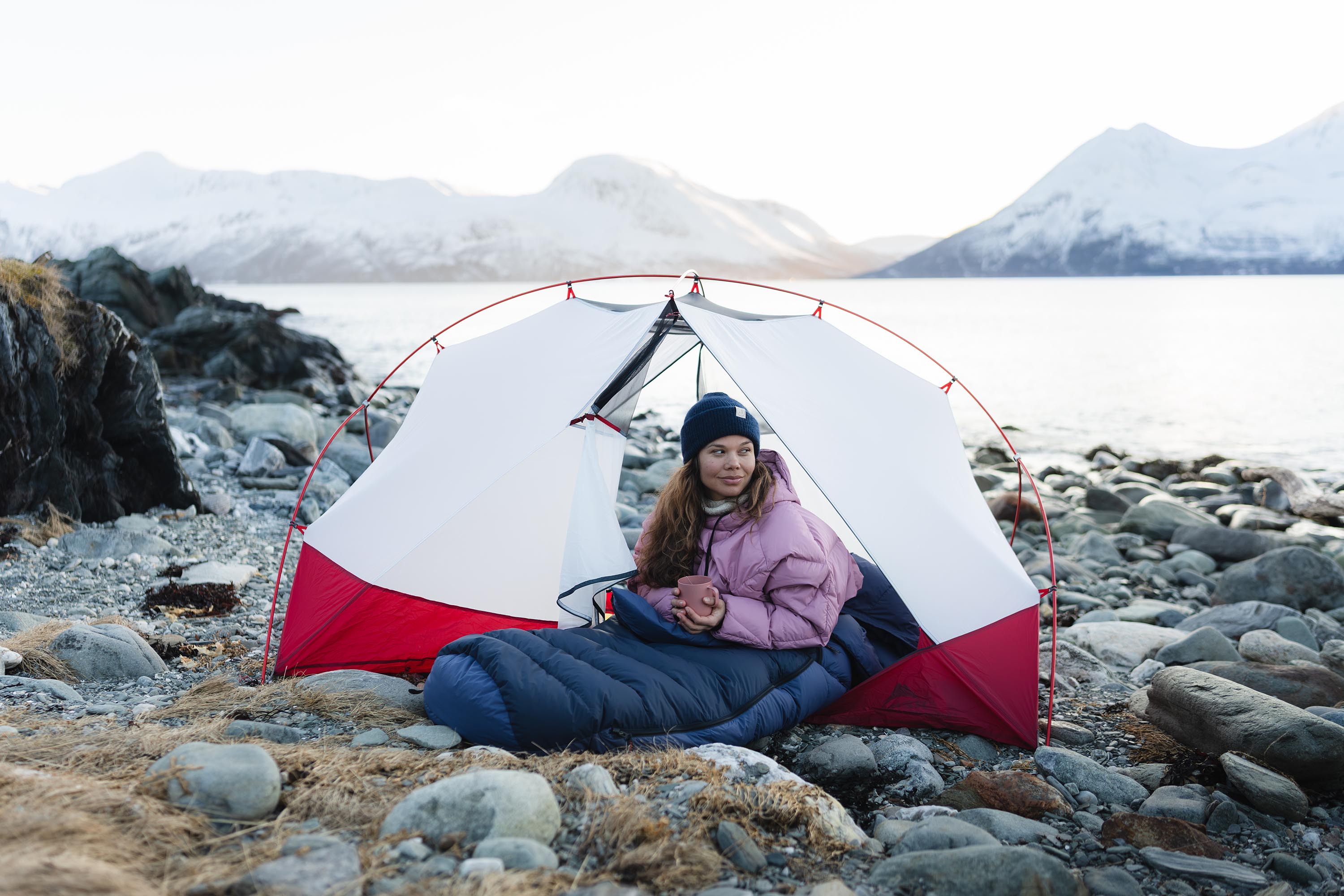 Person in a sleeping bag inside a red and white tent on a rocky beach with mountains in the background