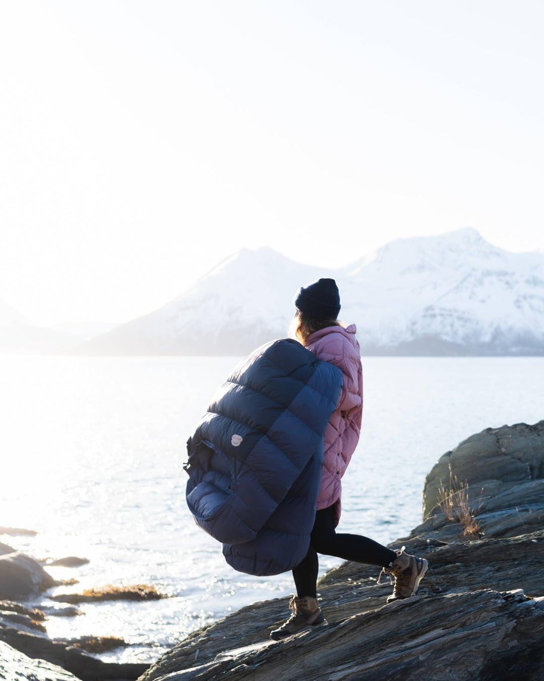 Person with a blue sleeping bag walking on rocky terrain near water