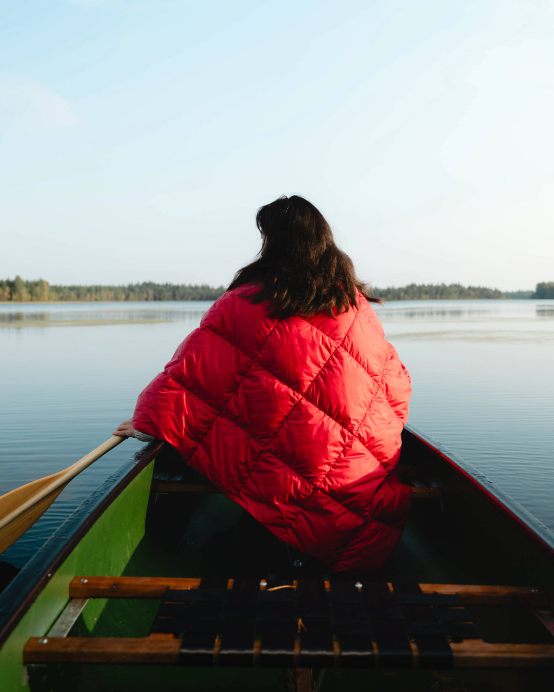 Person sitting in a canoe with their back to the camera, wearing a raspberry red Otava down poncho with diamond stitching. In the background, a calm lake and a tree-lined horizon.