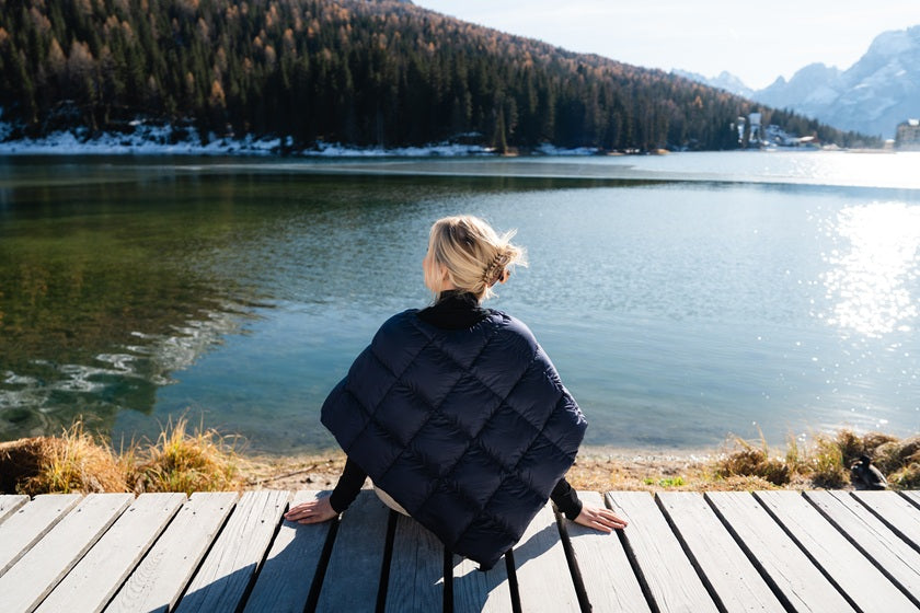 Person sitting on a wooden bench by a lake with mountains in the background. She is wearin dark blue Unelma down poncho