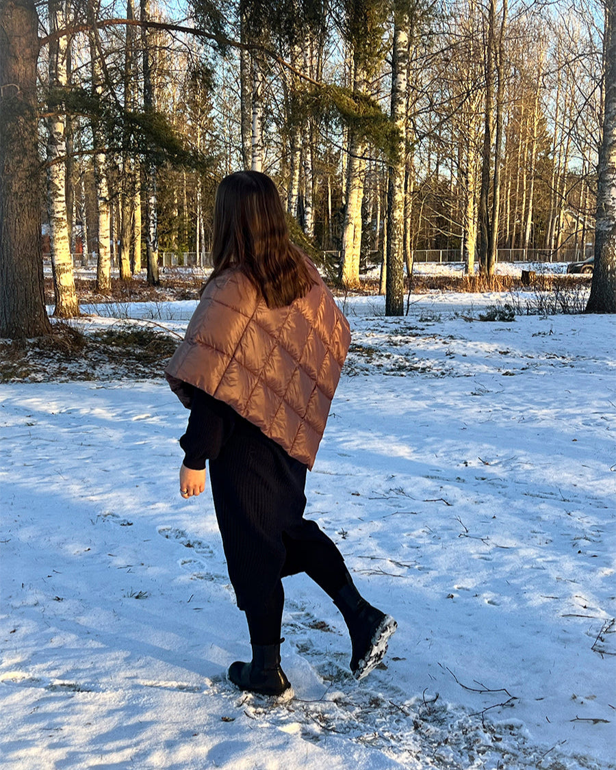A woman stands in a snowy forest landscape with her back to the camera, wearing a brown, diamond-quilted Unelma down poncho (color: hazelnut). The poncho covers her shoulders and upper back. She is wearing a black knitted dress and black winter boots.