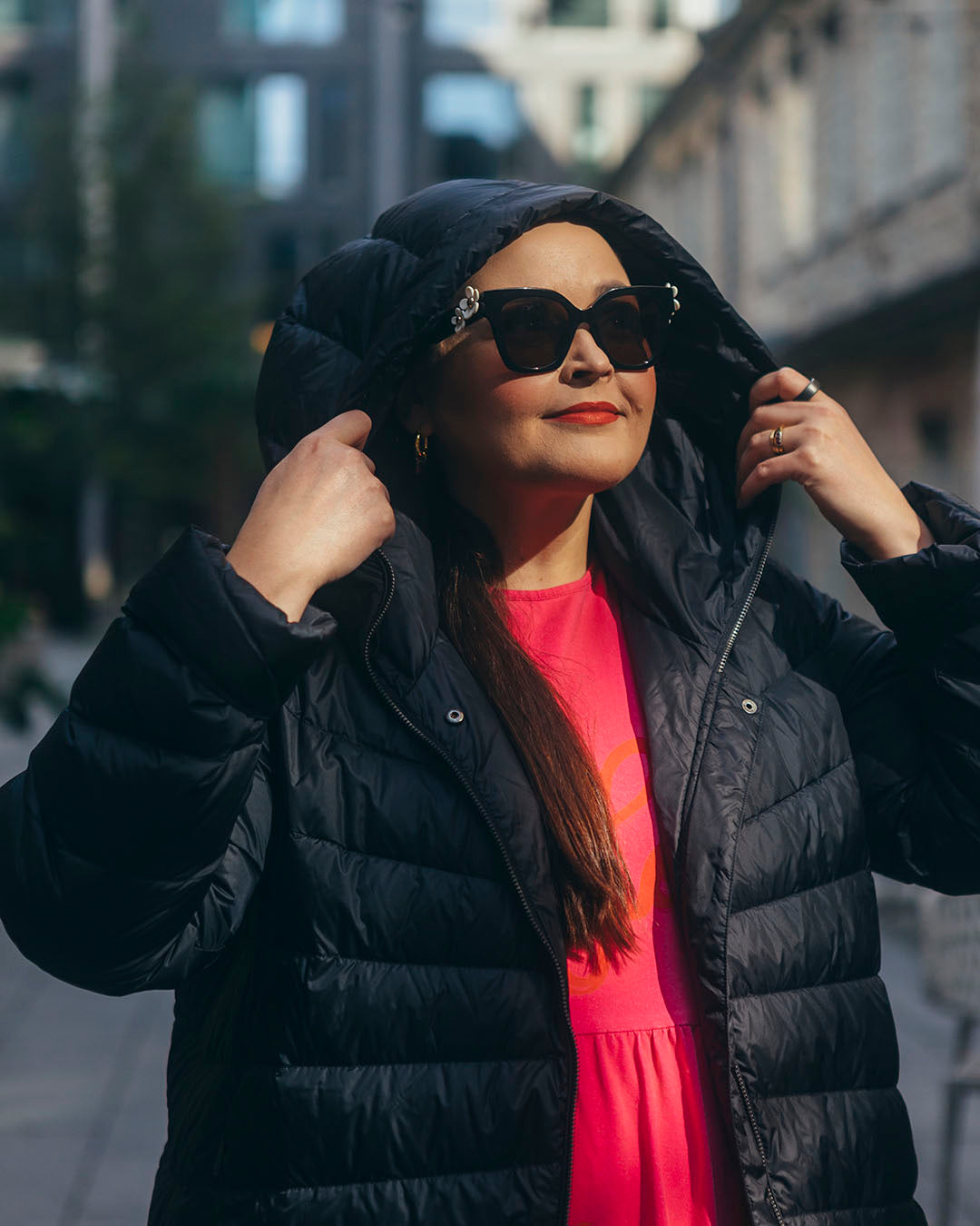 A woman is holding the hood of her black Säde light down coat (color: charcoal black) and wearing large sunglasses. The coat is open, revealing a bright pink dress underneath.