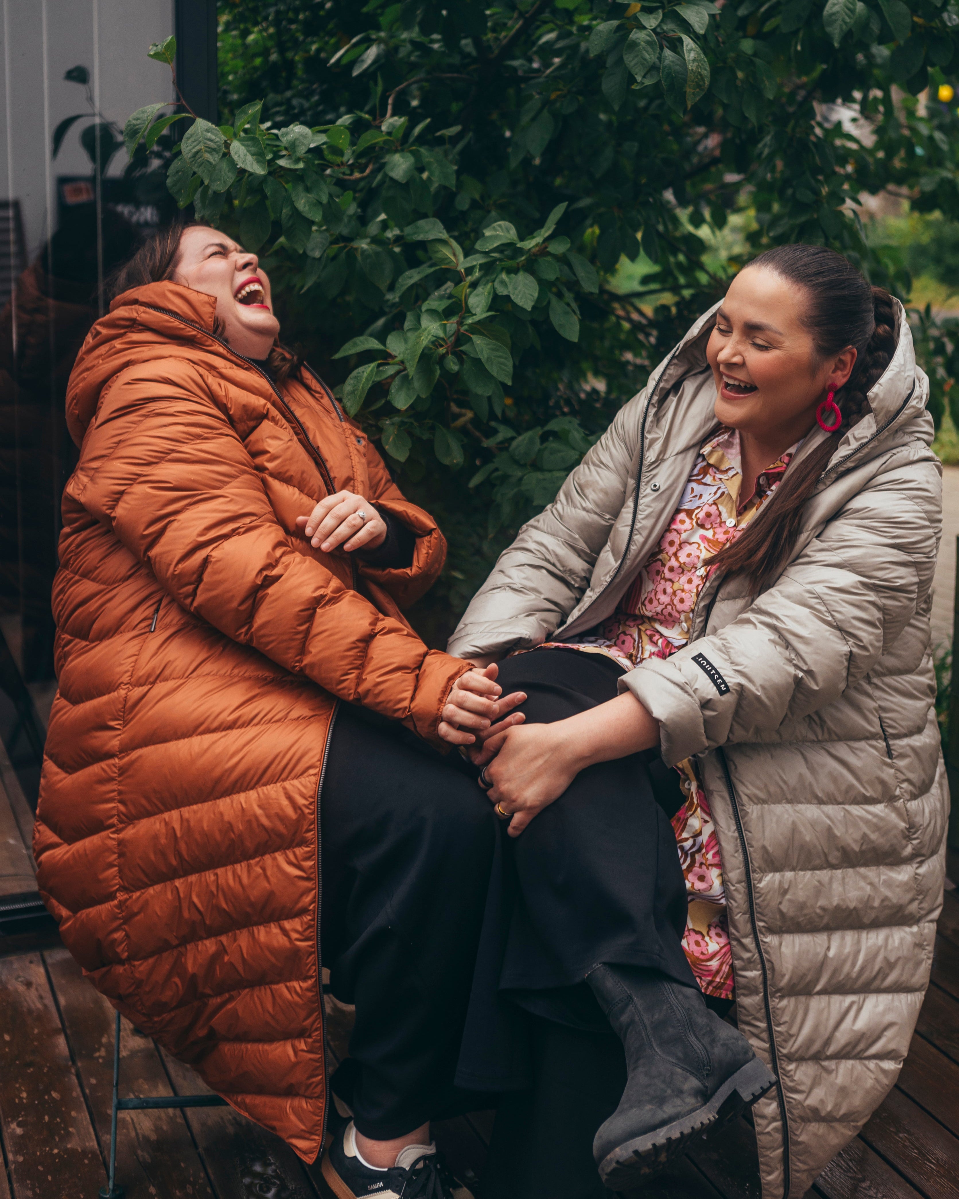 Two women are sitting side by side, laughing together. They are wearing muted orange (color: rose gold) and light (color: champagne) Säde light down coats, with black trousers visible underneath, and one of the women is wearing a colorful patterned dress.