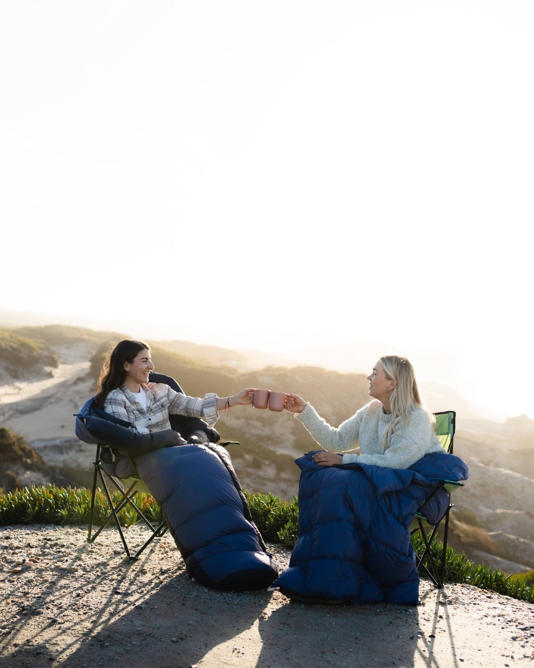Two people sitting in camping chairs with sleeping bags, holding coffee cups on a sandy beach.