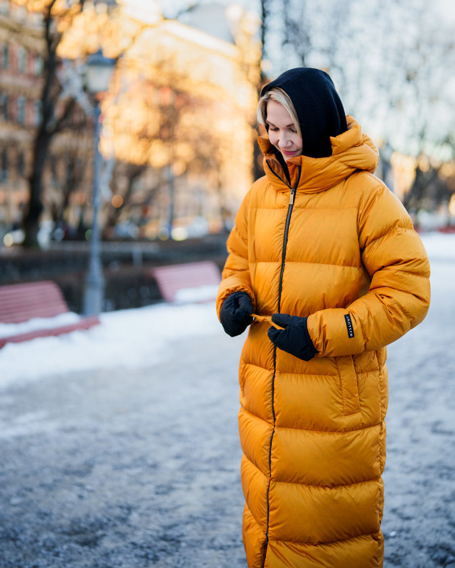 A woman stands on a winter street wearing an amber-colored Estrid down coat, black gloves, and a black hood. She is smiling and holding the drawstrings at the coat’s waist. In the background, there are benches, snow cover, and an urban landscape.