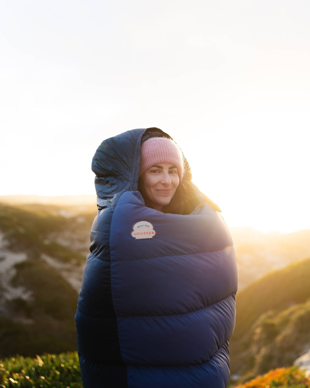 Person wearing a blue down sleeping bag with a brand logo, standing outdoors with a scenic background.