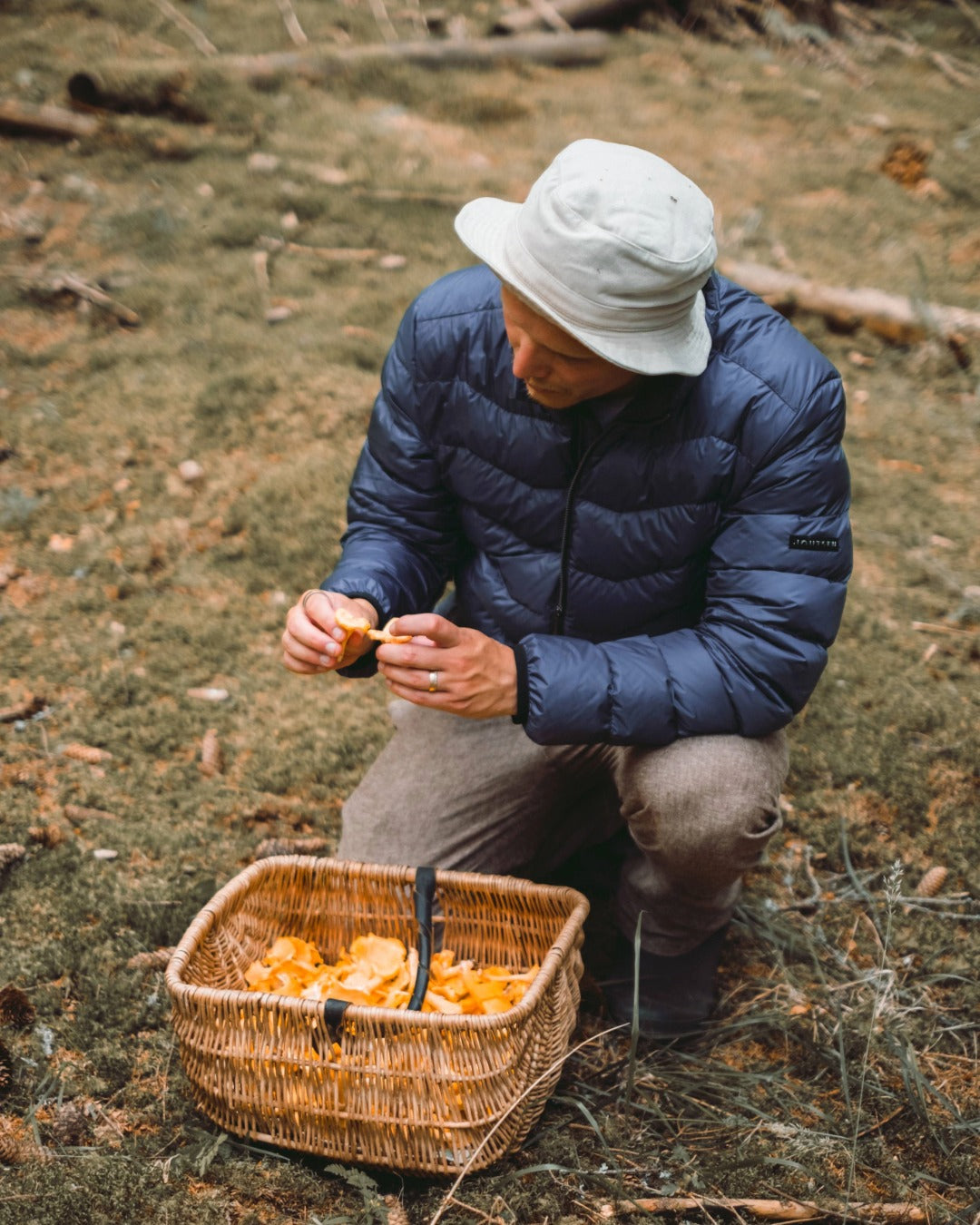 A man is crouching in the forest, cleaning chanterelles. He is wearing a blue Hile light down coat (color: Storm )