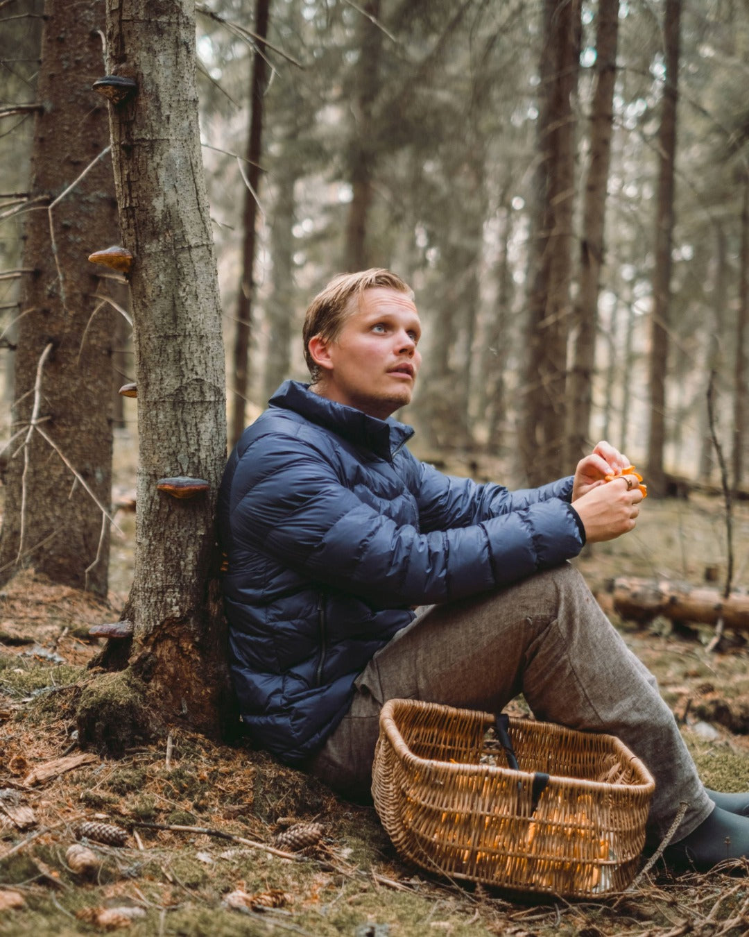 A man is sitting in the forest against a tree, holding mushrooms in his hands. He is wearing a blue Hile light down coat (color: Storm ), and in front of him is a woven basket.