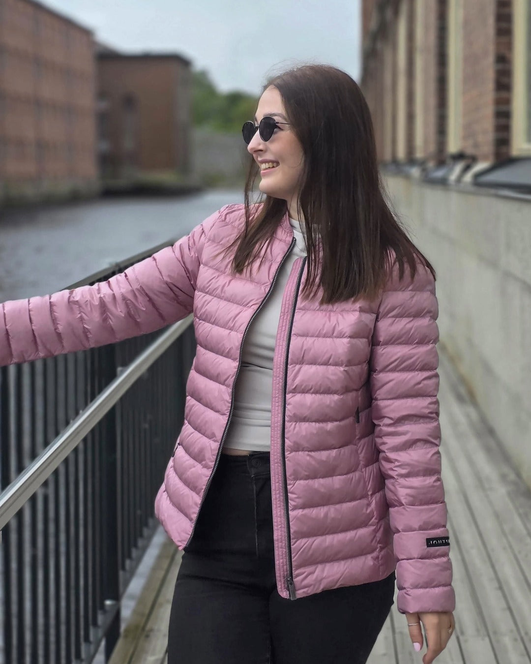 A woman is standing outside by a metal fence near the water. She is wearing a short, light pink Maana light down coat (color: Wildflower). The coat has horizontal quilting and zippered pockets.
