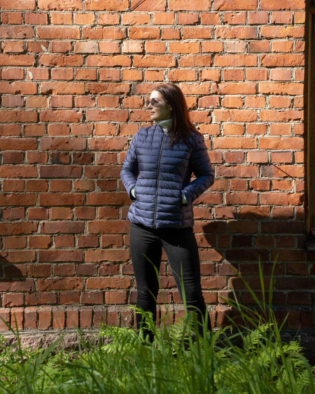 A woman stands in front of a brick wall with green plants in the foreground. She is wearing a blue Maana light down coat (color: Storm) and black trousers. The coat has zippered pockets and a small stand-up collar.