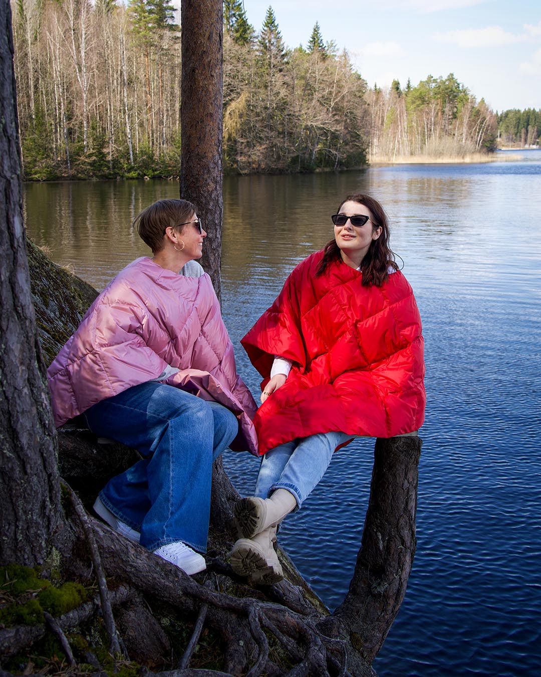 Two people sitting on tree roots by a lakeshore. The person on the left wears a light purple Otava down poncho (color: orchid), and the person on the right wears a red poncho (color: raspberry). Both are smiling and looking at each other.