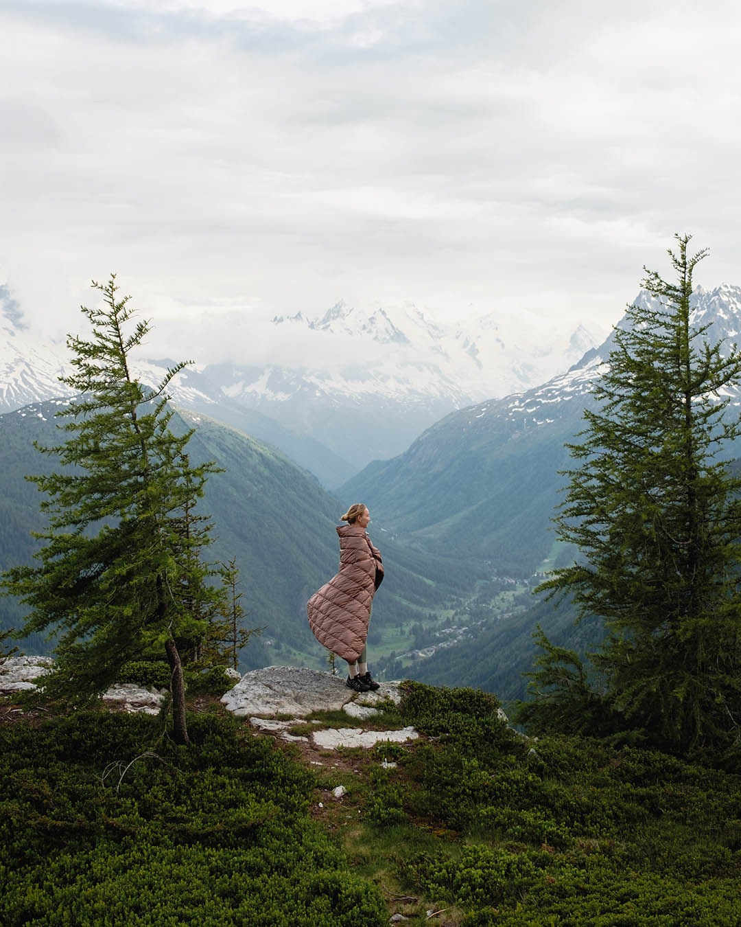 A person stands on a mountainside between two trees, looking out over the valley. They are dressed in a light brown Kulkuri down blanket (color: hazelnut) that billows in the wind. In the background, lush mountainous landscapes and snow-capped peaks are visible against a sky covered with clouds.
