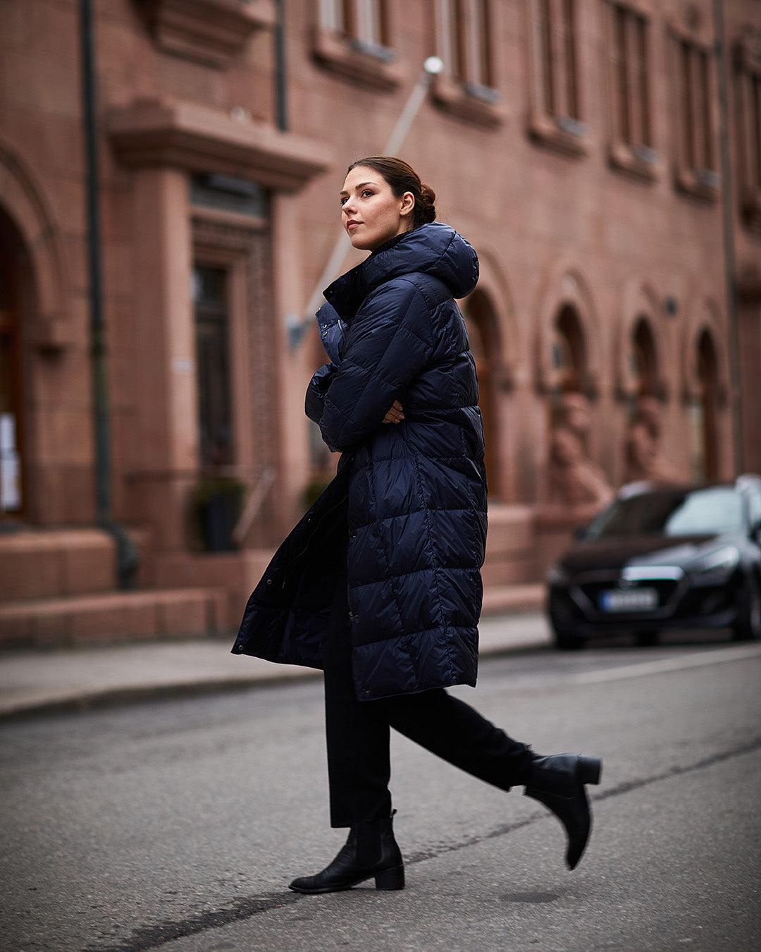 A woman walks along a city street wearing a dark blue, long Essi light down coat (color: Night Sky) with a hood and square quilting. Her outfit includes black pants and black ankle boots.