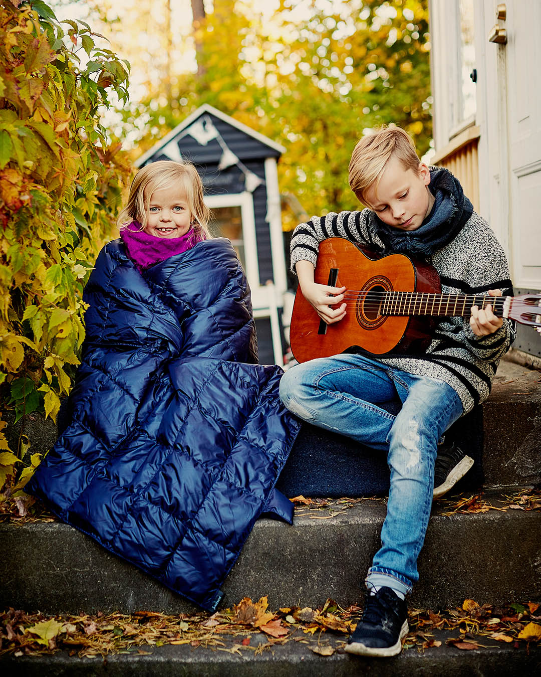 Two children are sitting on the steps. One child is wrapped in a dark blue Kulkuri down blanket and is smiling at the camera, while the other is wearing a grey wool sweater and a scarf, playing the guitar. In the background, colorful autumn bushes and a small cabin are visible.