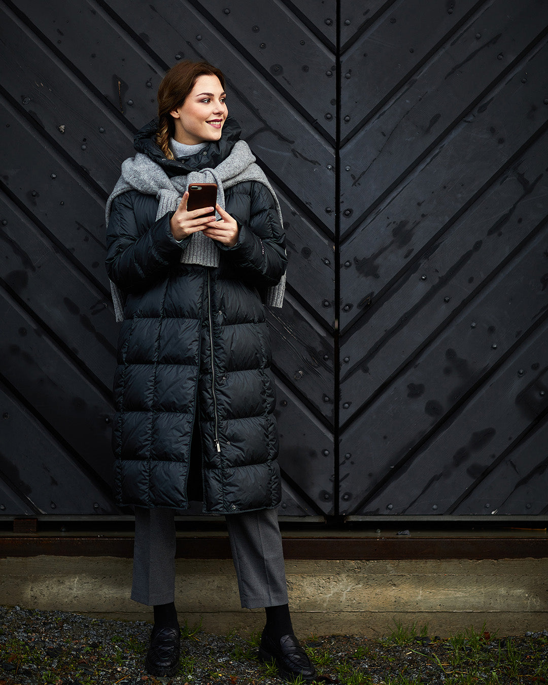 A woman wearing a black, long, slim-fit Essi light down coat (color: charcoal black) with square quilting. Her outfit includes a gray scarf, gray pants, and black shoes. She is holding a phone, and a black wooden door is visible in the background.