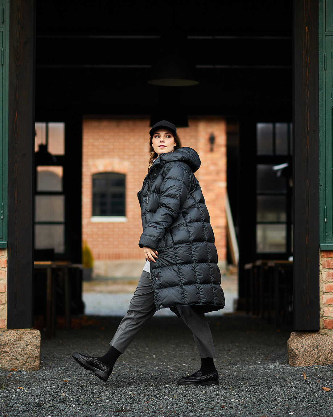 A woman wearing a black, long Essi light down coat (color: charcoal black) with a hood and square quilting. Her outfit includes gray pants, black shoes, and a black hat. A brick building is visible in the background.