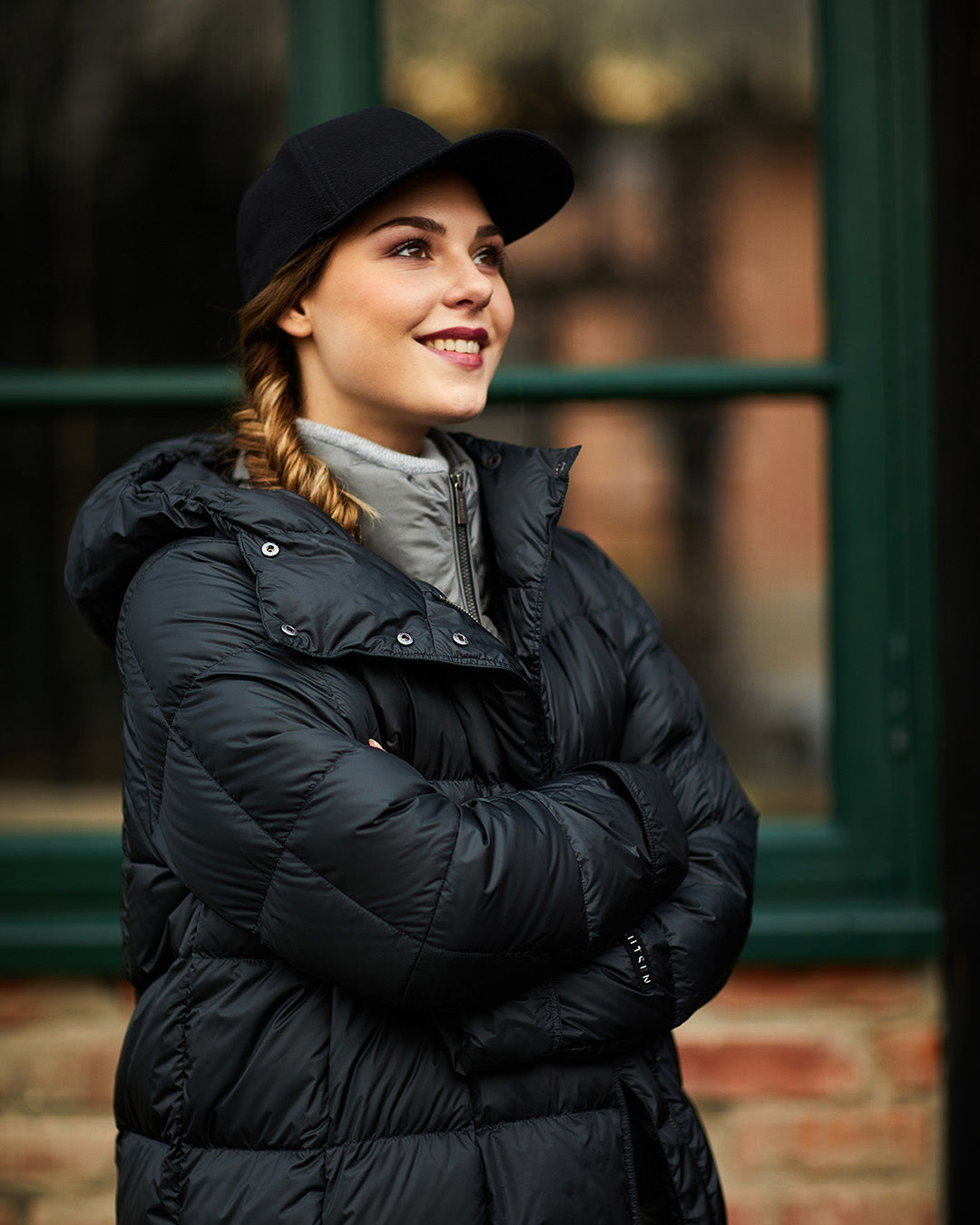 A woman wearing a black, thick, hooded Essi light down coat (color: charcoal black) with a light zip-up sweater underneath. She has a black baseball cap and braided hair. In the background, there is a green window frame and a brick wall.