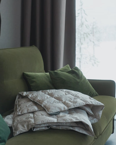 A woman is sitting on green sofa and cozying under a light (color: Seashell) Kulkuri down blanket.
