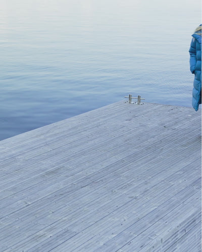 A blonde woman walking on a large pier wearing a blue (color name: Blue butterfly) long down coat. The coat’s zipper is open, and she has her hands in the coat pockets. She takes a few steps with a smile on her face.