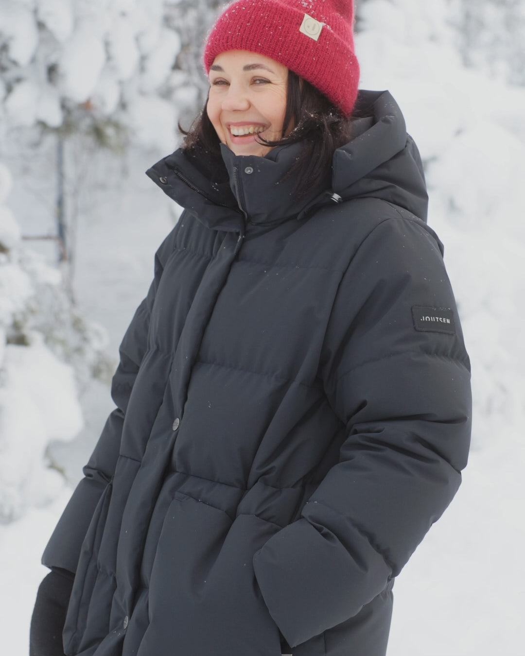 A women is wearing a black (color: jet black) down coat in a snowy landscape. She is wearing a red beanie.