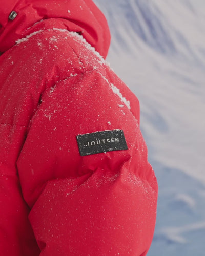 A man is standing in a snowy landscape, putting gloves to his pocket and closes the zipper. He is wearing a long, red (color: chili pepper) Tuisku down parka coat with a large hood. 