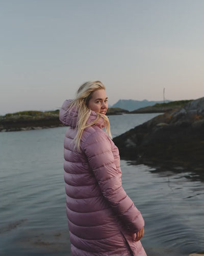 A woman stands by the seashore wearing a light pink Hanne light down coat (color: Wildflower) and puts her hands in the pockets.